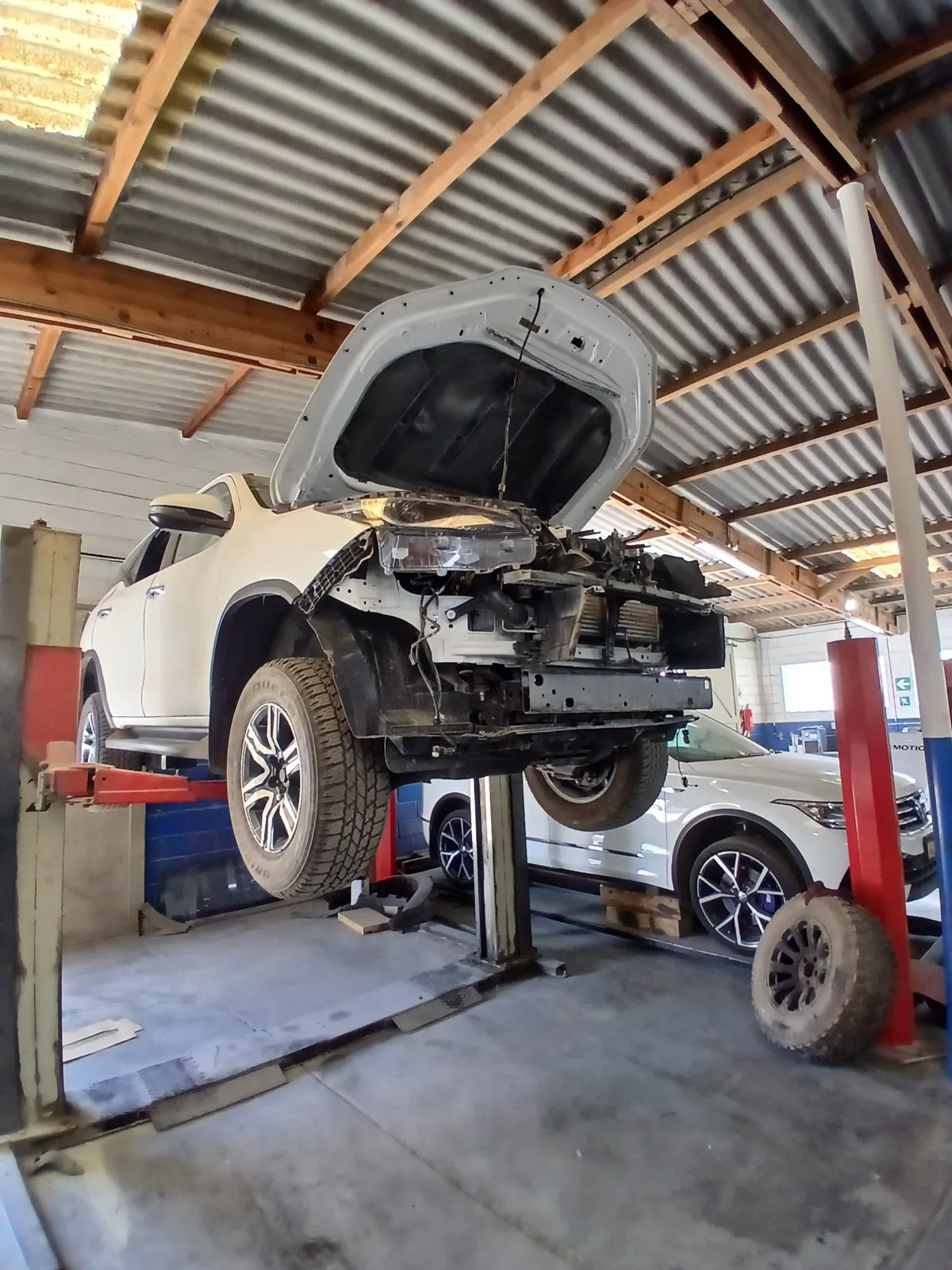 White SUV with front-end collision damage, stripped and raised on a lift in the Mototec workshop
